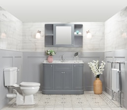 A well-organized bathroom features a gray vanity with a marble countertop, a round mirror, and a small shelf. Two wall sconces provide warm lighting. There is a pink vase with flowers on the vanity and a wicker basket with white flowers on the floor. A white toilet is positioned to the left, and a heated towel rail is on the right. The walls are tiled with a light marble pattern, and the floor has a light geometric design.