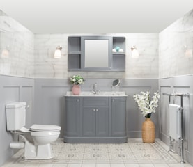 A well-organized bathroom features a gray vanity with a marble countertop, a round mirror, and a small shelf. Two wall sconces provide warm lighting. There is a pink vase with flowers on the vanity and a wicker basket with white flowers on the floor. A white toilet is positioned to the left, and a heated towel rail is on the right. The walls are tiled with a light marble pattern, and the floor has a light geometric design.