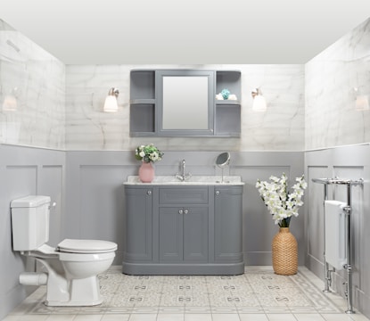 A well-organized bathroom features a gray vanity with a marble countertop, a round mirror, and a small shelf. Two wall sconces provide warm lighting. There is a pink vase with flowers on the vanity and a wicker basket with white flowers on the floor. A white toilet is positioned to the left, and a heated towel rail is on the right. The walls are tiled with a light marble pattern, and the floor has a light geometric design.