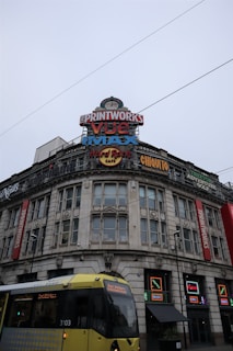 A large building with a vintage architectural style features multiple vibrant signs for entertainment venues, including The Printworks, Vue, IMAX, Hard Rock Cafe, and various restaurants. A yellow tram is visible in the foreground, moving past the building under an overcast sky.