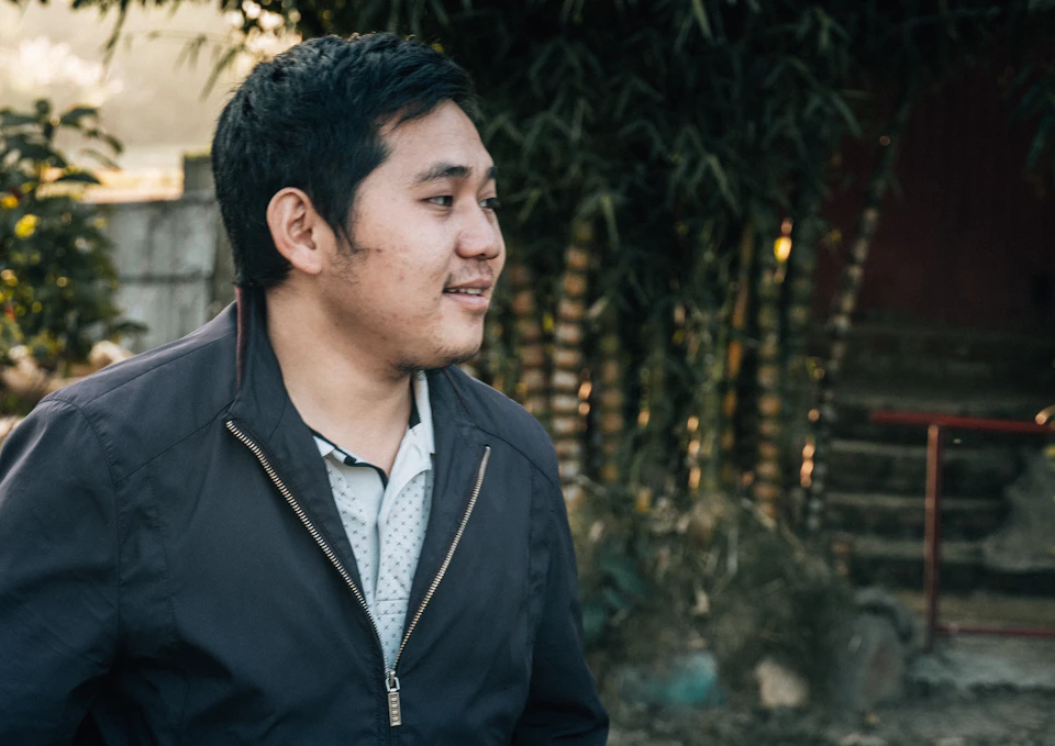 The image shows a man standing outdoors, wearing a black jacket and white shirt with blue accents. He is looking to his left side, possibly observing something or someone out of frame. The background features a natural setting with trees and bushes in various shades of green, creating a serene atmosphere. A red door can also be seen in the background, adding an interesting contrast to the greenery.