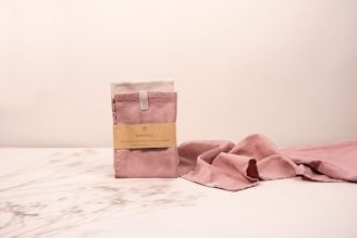 A set of pink cloth towels is neatly folded with a minimalist label attached, placed on a light surface. The cloth extends to the right in a gentle wave pattern. Soft, warm lighting casts gentle shadows, creating a relaxed atmosphere.