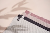 A set of three folded knit fabrics in varying colors with texture patterns. Each fabric has a small tag featuring a circular logo. The colors range from light gray to dark gray and soft pink. Shadows cast across the fabrics give a subtle sense of depth.