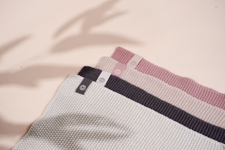 A set of three folded knit fabrics in varying colors with texture patterns. Each fabric has a small tag featuring a circular logo. The colors range from light gray to dark gray and soft pink. Shadows cast across the fabrics give a subtle sense of depth.