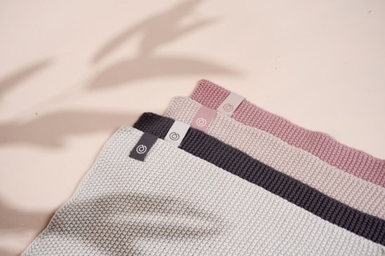 A set of three folded knit fabrics in varying colors with texture patterns. Each fabric has a small tag featuring a circular logo. The colors range from light gray to dark gray and soft pink. Shadows cast across the fabrics give a subtle sense of depth.