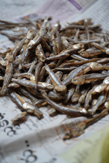 A pile of small dried fish is arranged on a sheet of newspaper. The fish are light brown in color with visible scales and eyes, indicating they have not been processed beyond drying.