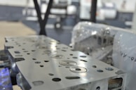A close-up view of a metal engine component with precise circular and rectangular cutouts. The surface is polished and reflects light, while in the background, there appears to be industrial machinery and protective wrapping.