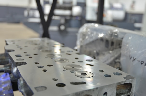 A close-up view of a metal engine component with precise circular and rectangular cutouts. The surface is polished and reflects light, while in the background, there appears to be industrial machinery and protective wrapping.