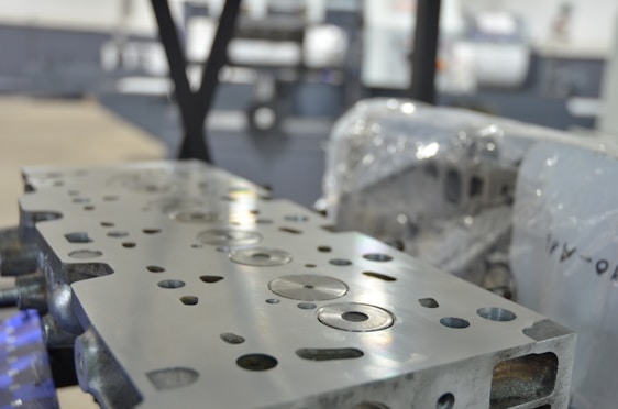 A close-up view of a metal engine component with precise circular and rectangular cutouts. The surface is polished and reflects light, while in the background, there appears to be industrial machinery and protective wrapping.