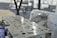 A close-up view of a metal engine component with precise circular and rectangular cutouts. The surface is polished and reflects light, while in the background, there appears to be industrial machinery and protective wrapping.