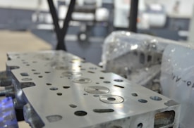 A close-up view of a metal engine component with precise circular and rectangular cutouts. The surface is polished and reflects light, while in the background, there appears to be industrial machinery and protective wrapping.