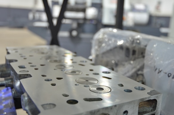 A close-up view of a metal engine component with precise circular and rectangular cutouts. The surface is polished and reflects light, while in the background, there appears to be industrial machinery and protective wrapping.