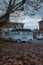A vintage van is parked on a street covered with fallen autumn leaves. The van has 'Nomad Kitchen & Bar' written on its side. The scene is set in an urban area with brick buildings in the background. A bare tree looms overhead, adding to the autumnal atmosphere.