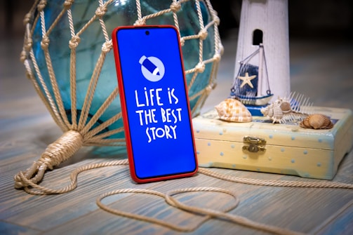 A smartphone encased in a red frame stands upright on a wooden surface. Its screen displays the text 'LIFE IS THE BEST STORY' with a simple graphic of a paper plane. The phone is positioned next to a nautical-themed decor, including a white wooden lighthouse figurine, a small wooden box with seashells on top, and a large, decorative glass buoy wrapped in knotted rope.