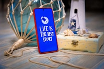 A smartphone encased in a red frame stands upright on a wooden surface. Its screen displays the text 'LIFE IS THE BEST STORY' with a simple graphic of a paper plane. The phone is positioned next to a nautical-themed decor, including a white wooden lighthouse figurine, a small wooden box with seashells on top, and a large, decorative glass buoy wrapped in knotted rope.