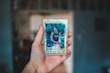 A hand is holding a basketball trading card encased in a protective plastic sleeve. The card features a player in a teal basketball jersey with the letters 'CHA' visible. The background is blurred, focusing attention on the foreground.
