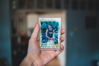 A hand is holding a basketball trading card encased in a protective plastic sleeve. The card features a player in a teal basketball jersey with the letters 'CHA' visible. The background is blurred, focusing attention on the foreground.
