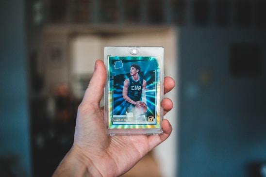 A hand is holding a basketball trading card encased in a protective plastic sleeve. The card features a player in a teal basketball jersey with the letters 'CHA' visible. The background is blurred, focusing attention on the foreground.