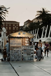 A small, illuminated souvenir stand is situated in a public area, likely a promenade with several palm trees and historic buildings in the background. The stand is filled with various souvenirs like mugs, magnets, and keychains. Several people, including a man with a stroller and a woman with a child, are browsing the items.