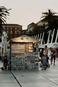 A small, illuminated souvenir stand is situated in a public area, likely a promenade with several palm trees and historic buildings in the background. The stand is filled with various souvenirs like mugs, magnets, and keychains. Several people, including a man with a stroller and a woman with a child, are browsing the items.