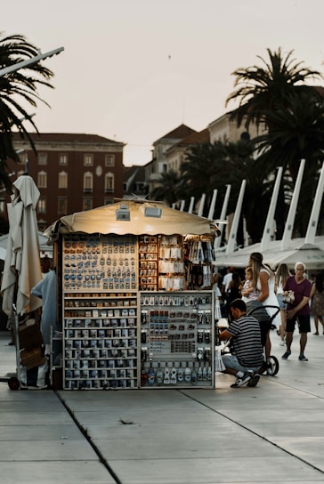A small, illuminated souvenir stand is situated in a public area, likely a promenade with several palm trees and historic buildings in the background. The stand is filled with various souvenirs like mugs, magnets, and keychains. Several people, including a man with a stroller and a woman with a child, are browsing the items.