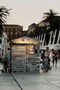 A small, illuminated souvenir stand is situated in a public area, likely a promenade with several palm trees and historic buildings in the background. The stand is filled with various souvenirs like mugs, magnets, and keychains. Several people, including a man with a stroller and a woman with a child, are browsing the items.