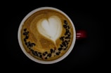 A cup of coffee with creamy latte art in the shape of a heart at its center. The top surface is decorated with a semi-circle of dark coffee beans or chocolate pieces. The cup has a red handle, and the background is black.