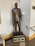 A bronze statue of a man in a suit holding a briefcase, placed on a pedestal with an inscription. The statue is located indoors near a corner with a wall-mounted lamp above it and a railing to the side.