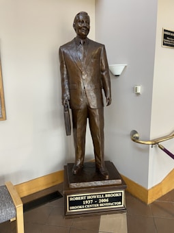 A bronze statue of a man in a suit holding a briefcase, placed on a pedestal with an inscription. The statue is located indoors near a corner with a wall-mounted lamp above it and a railing to the side.