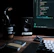 A dimly lit workspace with a computer monitor displaying code, a speaker, headphones, a mug with the text 'Develop. Preview. Ship.', and a notebook labeled 'Field Notes' on a wooden desk. The keyboard and a mouse are also visible.