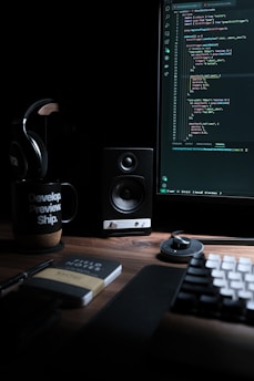 A dimly lit workspace with a computer monitor displaying code, a speaker, headphones, a mug with the text 'Develop. Preview. Ship.', and a notebook labeled 'Field Notes' on a wooden desk. The keyboard and a mouse are also visible.