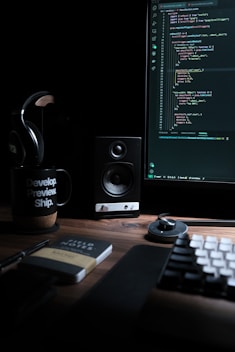 A dimly lit workspace with a computer monitor displaying code, a speaker, headphones, a mug with the text 'Develop. Preview. Ship.', and a notebook labeled 'Field Notes' on a wooden desk. The keyboard and a mouse are also visible.