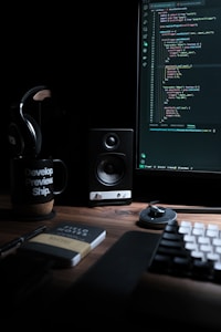 A dimly lit workspace with a computer monitor displaying code, a speaker, headphones, a mug with the text 'Develop. Preview. Ship.', and a notebook labeled 'Field Notes' on a wooden desk. The keyboard and a mouse are also visible.