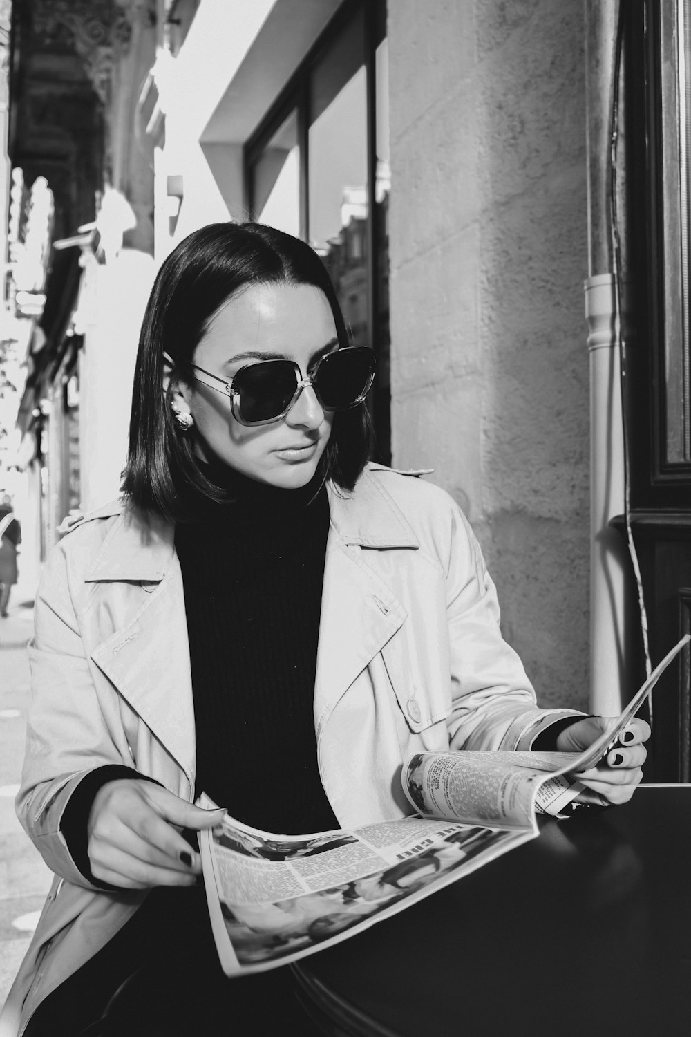 a woman in a trench coat and sunglasses reading a newspaper