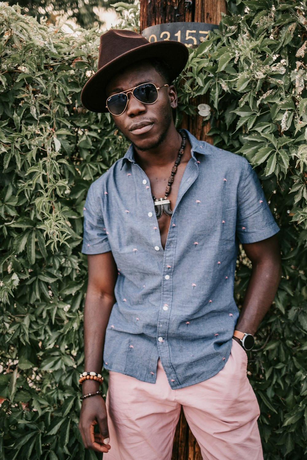 a man wearing a hat and sunglasses standing in front of a bush