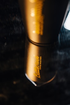 A sleek, metallic coffee tumbler with the branding 'Dante's Coffee Roasters' in elegant yellow text, reflecting on a dark, glossy surface. The lighting highlights the luxurious gold finish of the tumbler.
