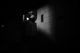 A dimly lit, monochromatic architectural structure with a strong focus on shadows and light. A singular light source illuminates a section of the building, creating a dramatic contrast against the surrounding darkness. The building features a window and geometric patterns, highlighting an urban, minimalist design.