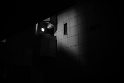 A dimly lit, monochromatic architectural structure with a strong focus on shadows and light. A singular light source illuminates a section of the building, creating a dramatic contrast against the surrounding darkness. The building features a window and geometric patterns, highlighting an urban, minimalist design.