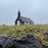 A small black church, slightly blurred in the background, is set against a grey, overcast sky. In the foreground, there are sharp details of grass and a rugged rock. The perspective emphasizes the natural elements more than the church.