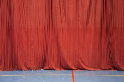 A long red curtain extends from ceiling to floor, creating a rich and dramatic backdrop. The surface of the curtain has subtle folds and drapes that give it texture. The floor beneath is a smooth blue with two painted lines, one red and one yellow, suggesting a sports or auditorium setting.
