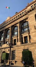 A grand, historic building adorned with classical architectural details. Notable features include arched windows, columns, and intricate stone carvings. A flagpole with the Mexican flag is visible atop the building. The structure is flanked by well-trimmed green bushes and a couple of people are seen walking in front.