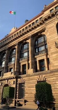 A grand, historic building adorned with classical architectural details. Notable features include arched windows, columns, and intricate stone carvings. A flagpole with the Mexican flag is visible atop the building. The structure is flanked by well-trimmed green bushes and a couple of people are seen walking in front.