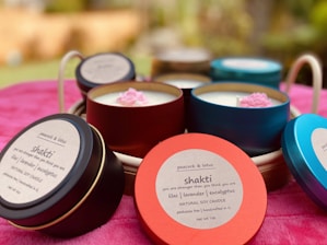 Several colorful candle tins are arranged on a soft, pink surface. The tins are labeled with 'peacock & lotus' and 'shakti,' indicating they are natural soy candles with lilac, lavender, and eucalyptus scents. Each tin has a prominent lid, and small decorative flowers are visible on the candle surfaces.