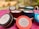Several colorful candle tins are arranged on a soft, pink surface. The tins are labeled with 'peacock & lotus' and 'shakti,' indicating they are natural soy candles with lilac, lavender, and eucalyptus scents. Each tin has a prominent lid, and small decorative flowers are visible on the candle surfaces.