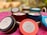 Several colorful candle tins are arranged on a soft, pink surface. The tins are labeled with 'peacock & lotus' and 'shakti,' indicating they are natural soy candles with lilac, lavender, and eucalyptus scents. Each tin has a prominent lid, and small decorative flowers are visible on the candle surfaces.