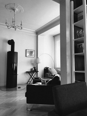 A black and white image of a modern living room with a cylindrical stove, a chandelier on the ceiling, a comfortable chair, a coffee table with a lamp and vase, and a cozy sofa. Shelving with glasses and books is visible, and there is parquet flooring.