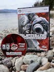 A PC DVD game and its disc are placed on a rocky shoreline. The game box features futuristic, industrial-themed artwork with gears and mechanical elements, while the disc shares a similar design. The background features a body of water and blurred trees, suggesting an outdoor setting.