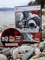 A PC DVD game and its disc are placed on a rocky shoreline. The game box features futuristic, industrial-themed artwork with gears and mechanical elements, while the disc shares a similar design. The background features a body of water and blurred trees, suggesting an outdoor setting.
