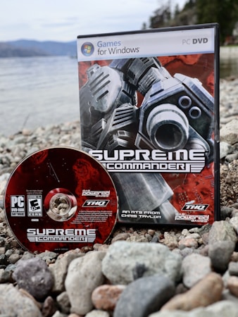 A PC DVD game and its disc are placed on a rocky shoreline. The game box features futuristic, industrial-themed artwork with gears and mechanical elements, while the disc shares a similar design. The background features a body of water and blurred trees, suggesting an outdoor setting.