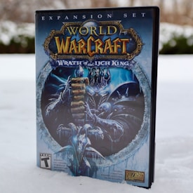 A video game box for the expansion set 'World of Warcraft: Wrath of the Lich King' is placed upright on snowy ground. The cover features an imposing armored figure, holding a large sword, with icy blue and white color themes. The Blizzard Entertainment logo is visible at the bottom along with other game information.
